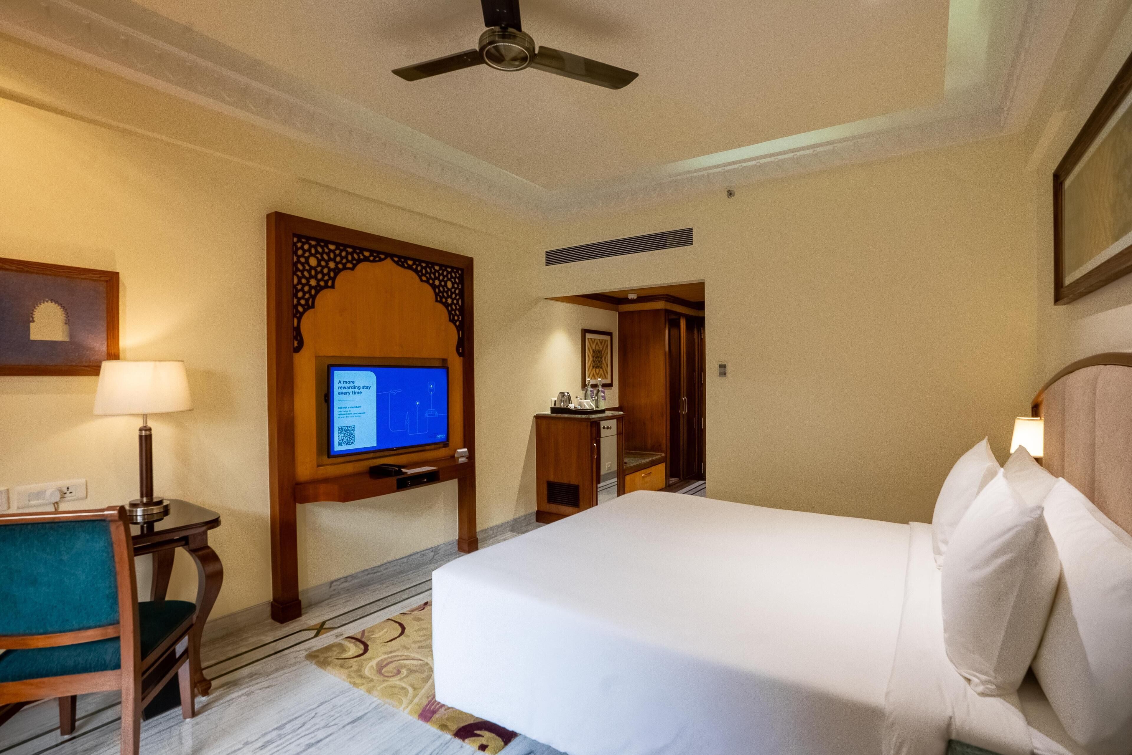 A hotel room with a bed, a desk with a computer, a chair, and a ceiling fan.
