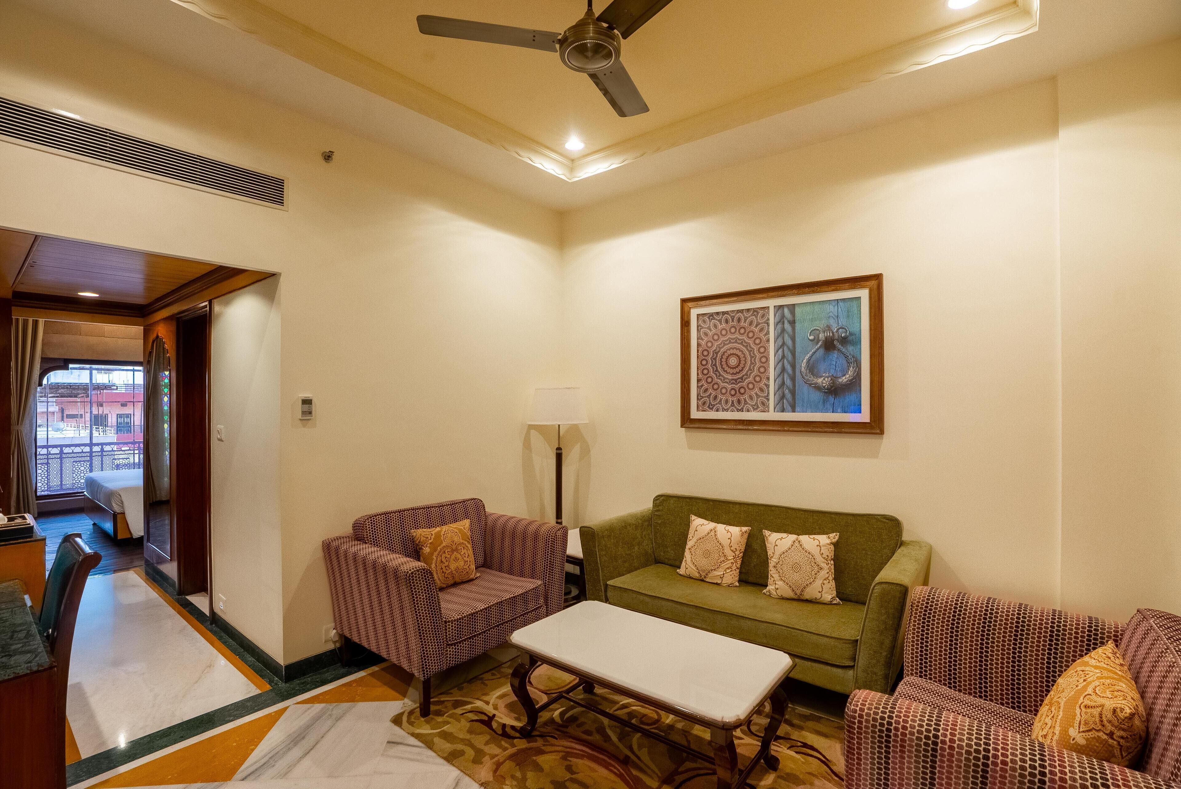 A hotel room with a green sofa, a patterned armchair, a white coffee table, and a ceiling fan.