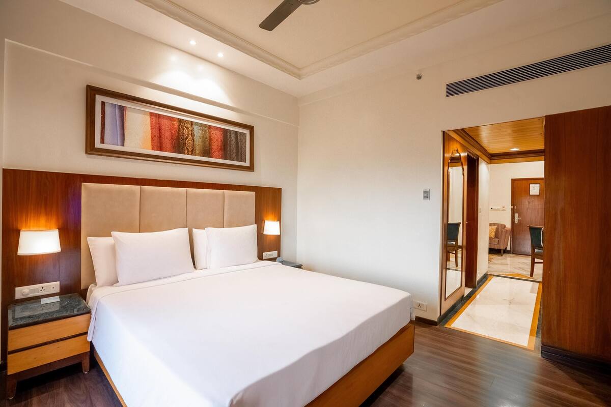 Ein Hotelzimmer mit einem großen Bett, einem Holzkopfende, Nachttischlampen, einem gerahmten Kunstwerk an der Wand und einer Holztür, die in einen anderen Raum führt.
