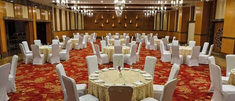 Un salón de banquetes con mesas redondas dispuestas para comer, sillas blancas, candelabros y un suelo cubierto de alfombra roja.