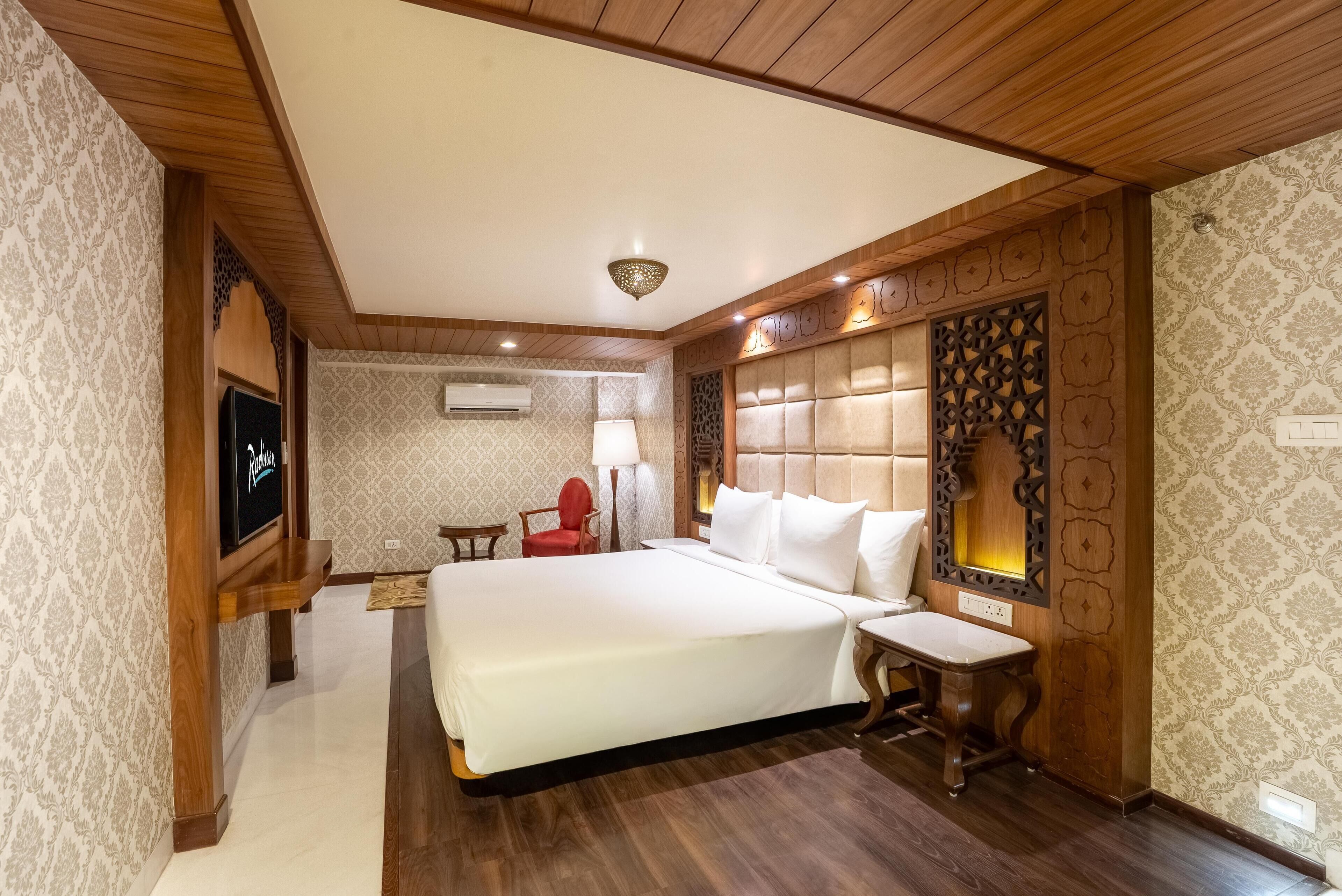 A hotel room with a large bed, a TV mounted on the wall, a red chair, and a decorative wall panel.