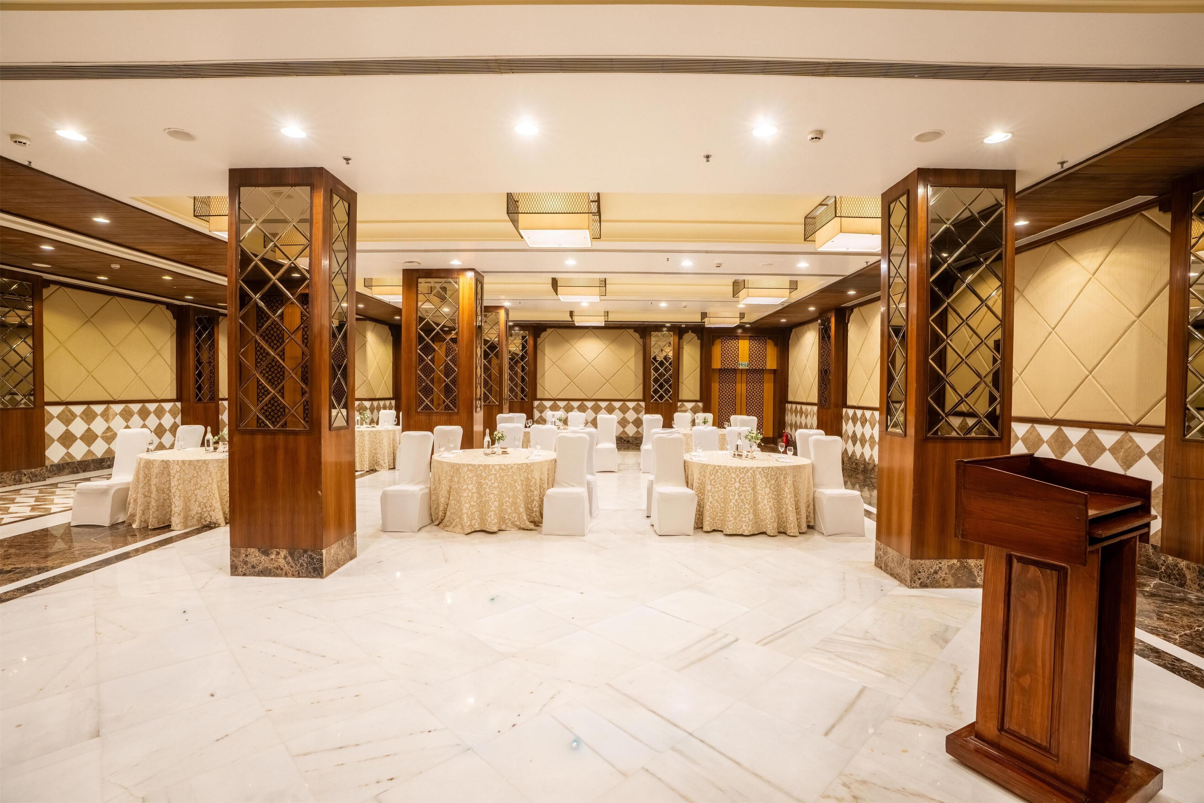 Un amplio salón de banquetes con mesas redondas y sillas blancas, pilares de madera y suelo de mármol.