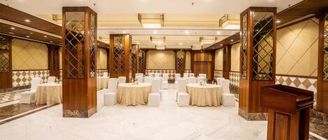 Un amplio salón de banquetes con mesas redondas y sillas blancas, pilares de madera y suelo de mármol.
