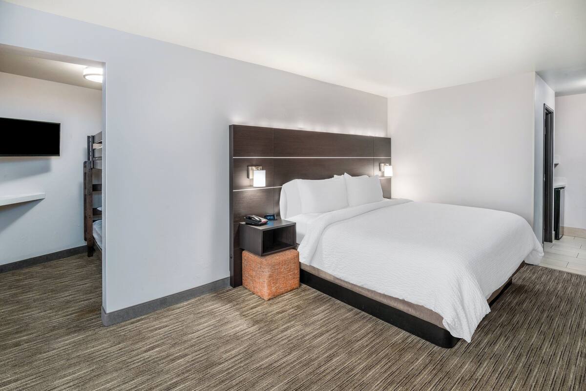 A modern hotel room with a large bed, a bedside table with a lamp, a flat-screen TV mounted on the wall, and a small wooden side table.