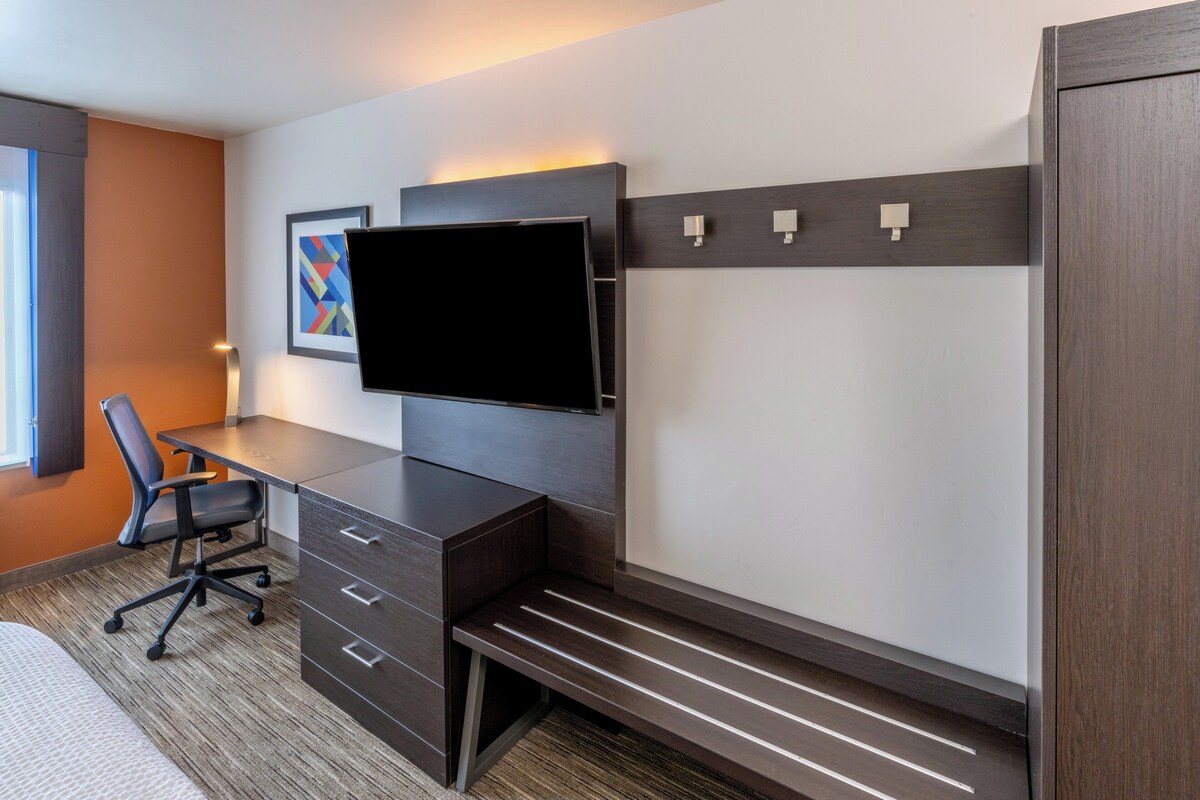 Quarto de hotel moderno com escrivaninha, cadeira, televisão e espaço de armazenamento embutido.