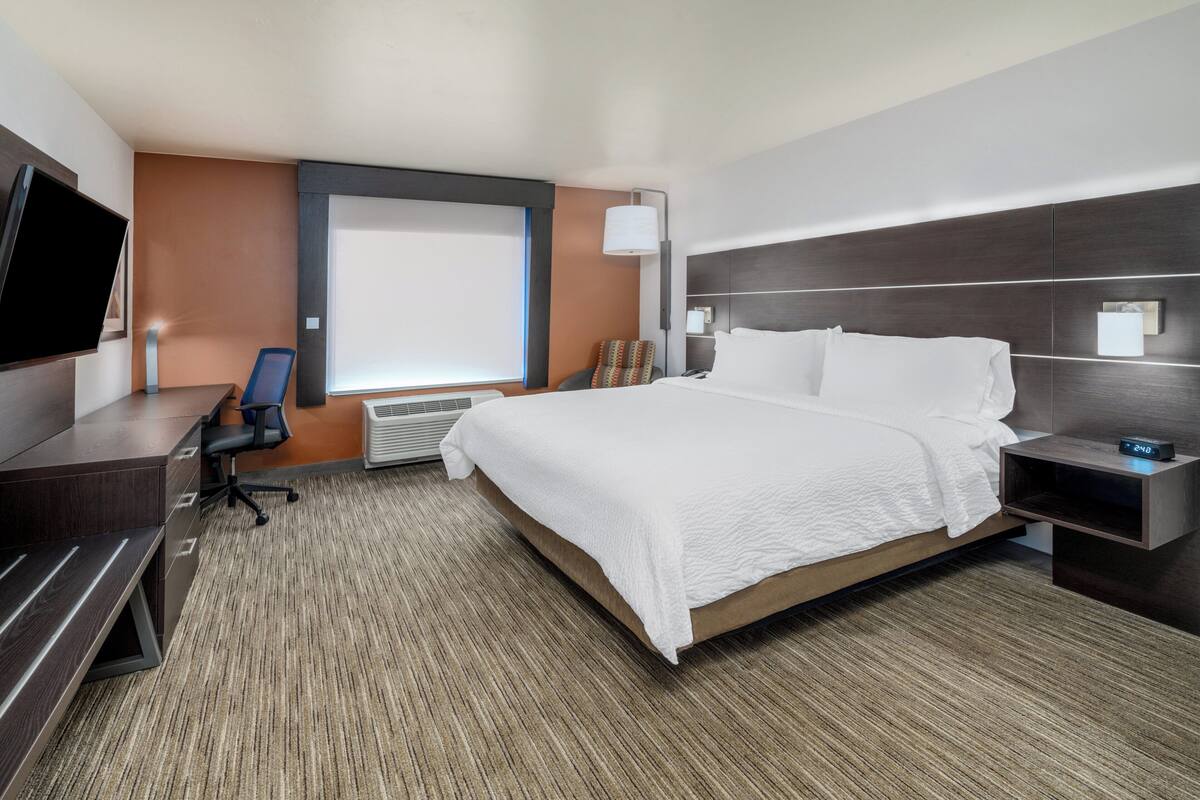 A hotel room with a large bed, a desk with a chair, a flat-screen TV, and a window with blinds.