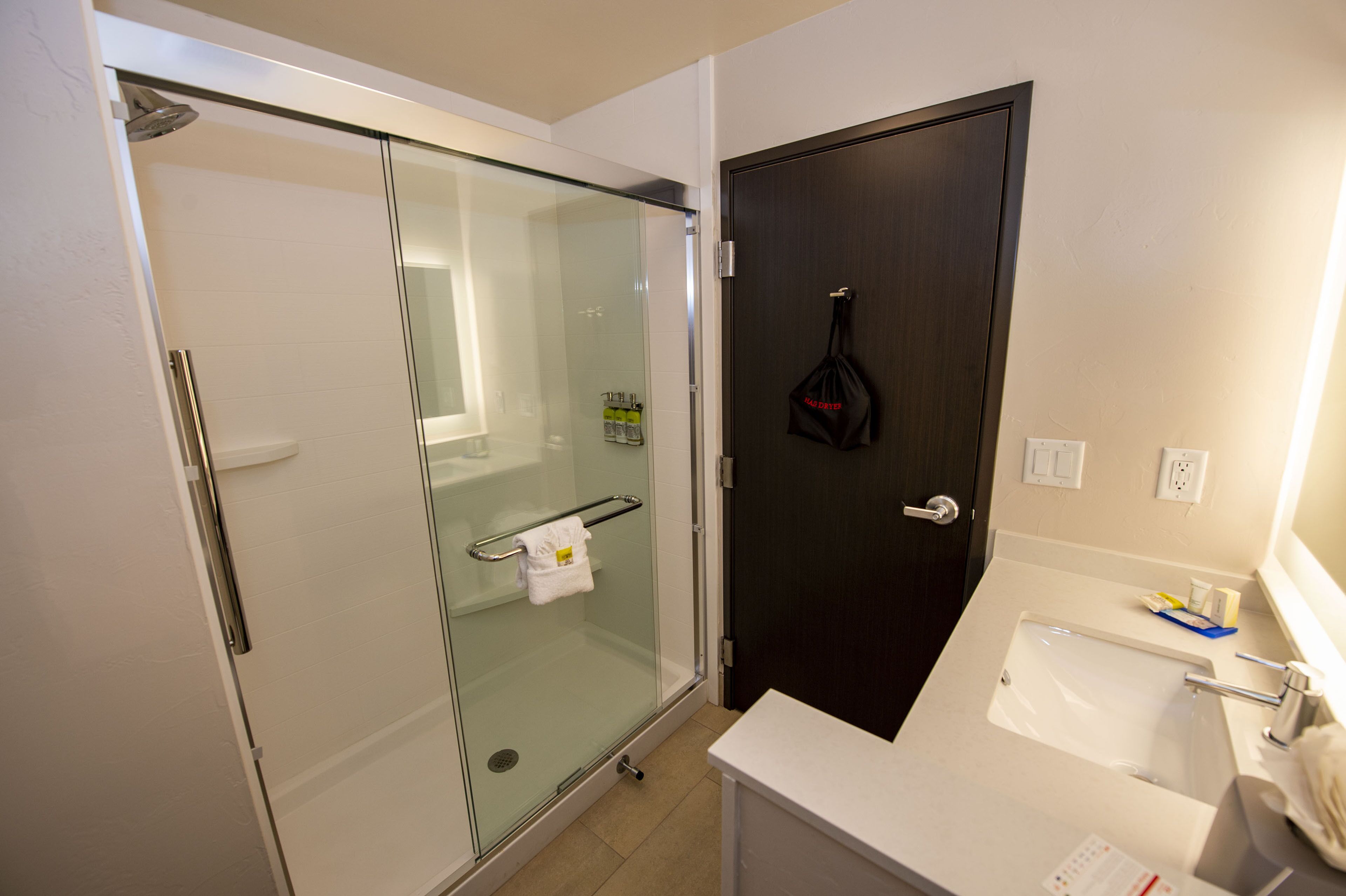A modern bathroom with a glass-enclosed shower, a dark wooden door, and a white sink with toiletries.