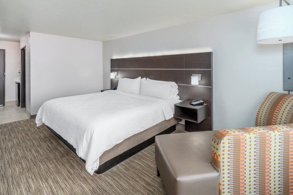 Quarto de hotel moderno com uma cama grande, uma mesinha de cabeceira, uma cadeira e um abajur.