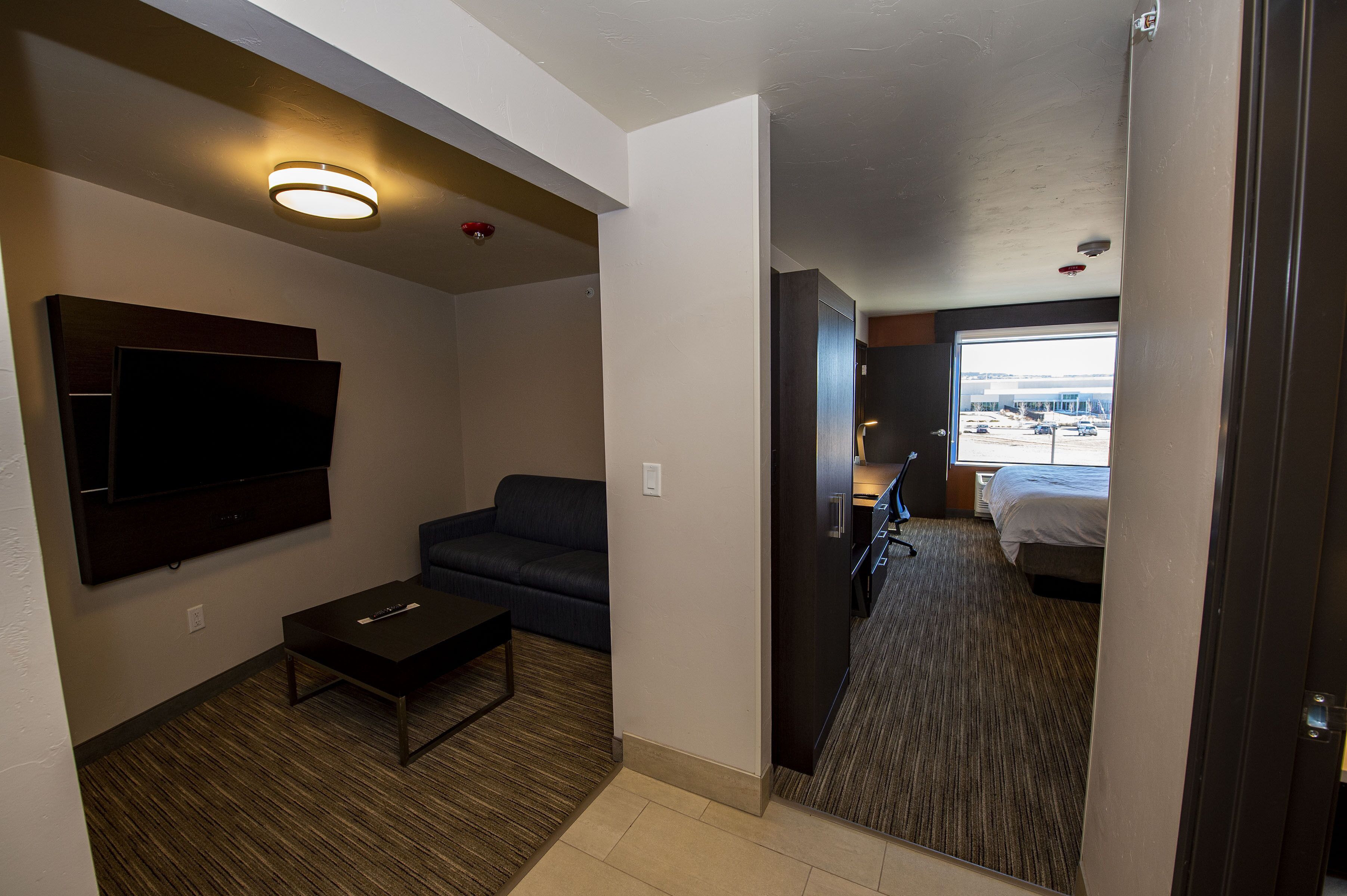 A hotel room with a flat-screen TV mounted on the wall, a dark-colored sofa, a small coffee table, and a bed with a view of the outside.