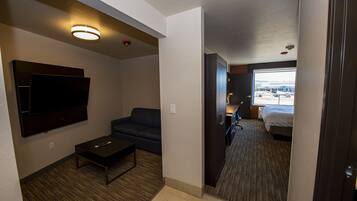 A hotel room with a flat-screen TV mounted on the wall, a dark-colored sofa, a small coffee table, and a bed with a view of the outside.