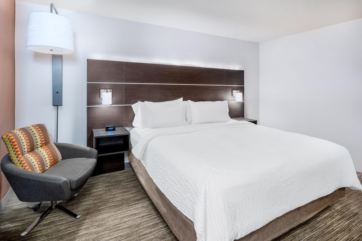 A modern hotel room with a large bed, a gray armchair, a bedside table with a lamp, and a wall-mounted shelf.