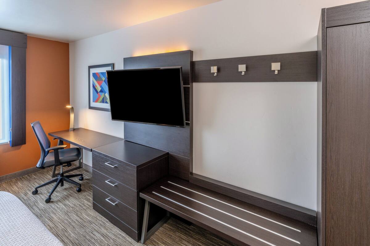 A modern hotel room with a desk, chair, TV, and built-in storage.