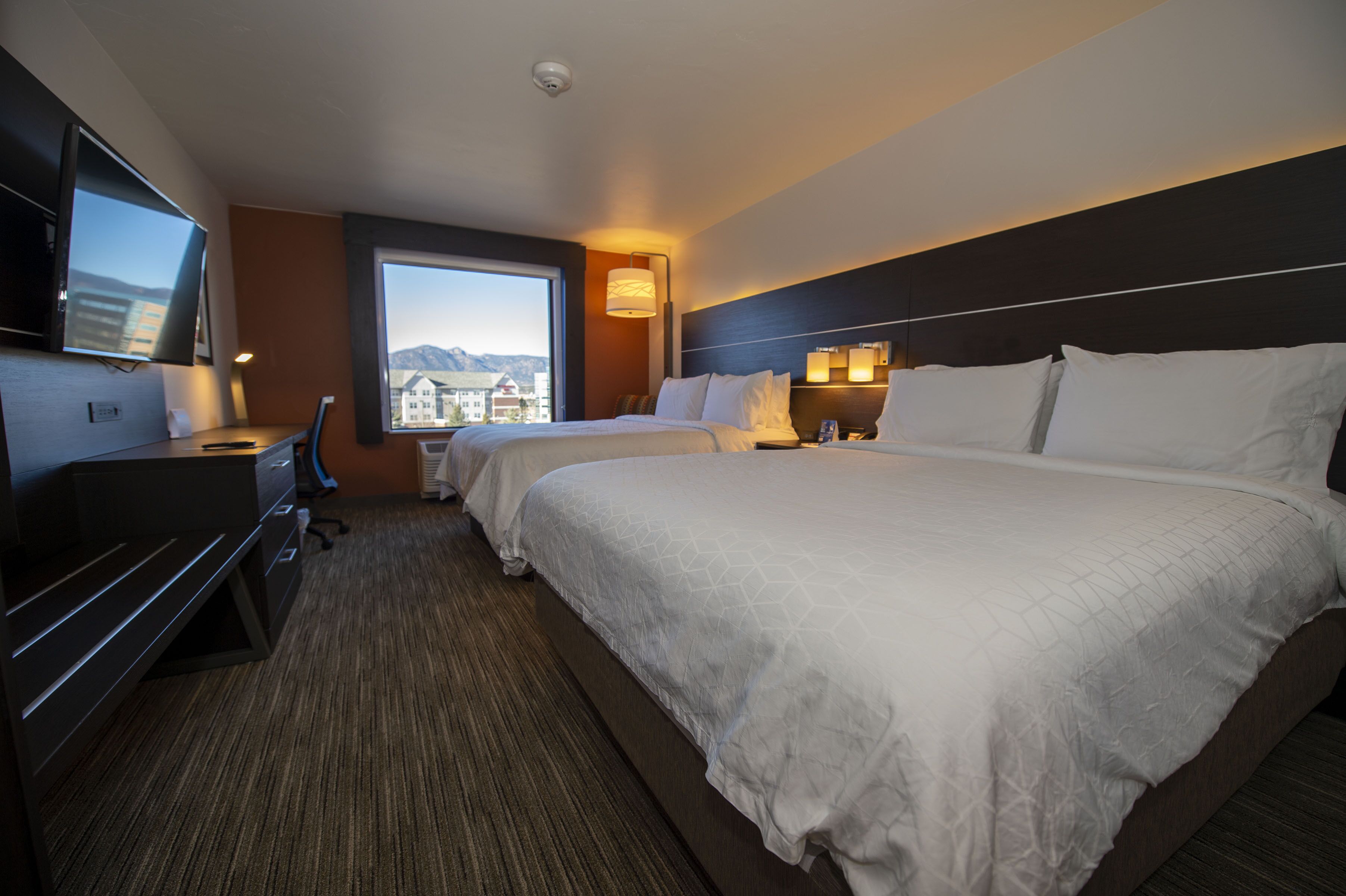 Une chambre d’hôtel avec deux lits, un bureau, une télévision et une fenêtre donnant sur des immeubles et des montagnes.