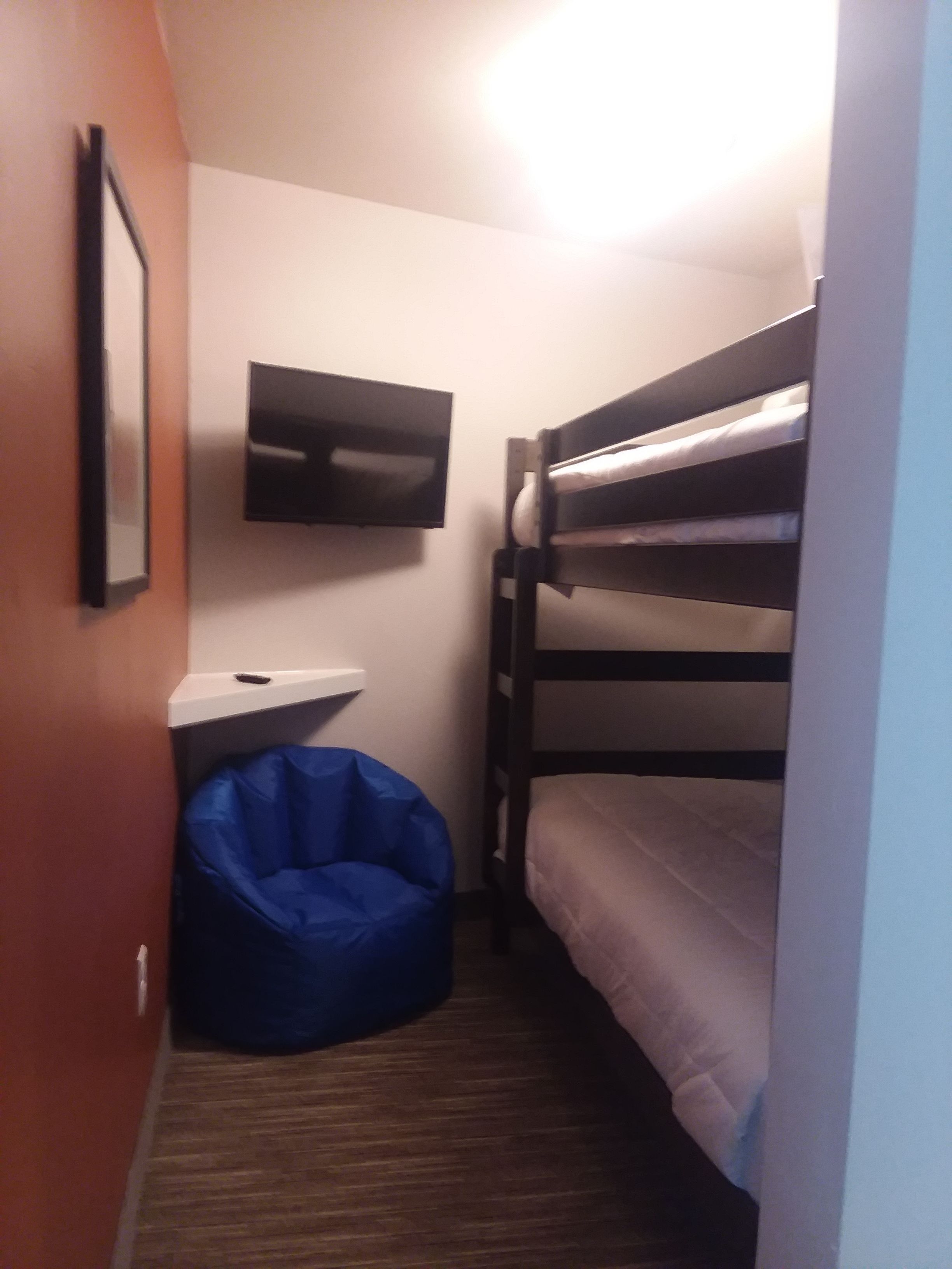 A small, narrow room with a bunk bed, a flat-screen TV mounted on the wall, and a blue bean bag chair.