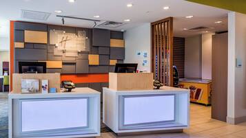 Área de recepção com design moderno, incluindo balcão de check-in, display digital e placa com o nome do hotel.
