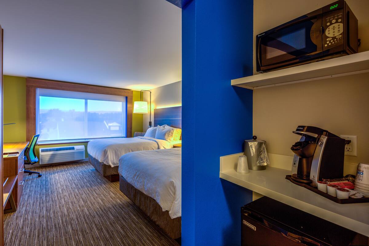A hotel room with two beds, a desk with a computer, a coffee machine, and a view of the outdoors.