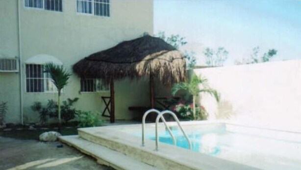 A swimming pool with a thatched roof structure, a concrete deck, and a building in the background. - Casa en PlayaDelCarmen 510 by Sinbad (Playa del Carmen)
