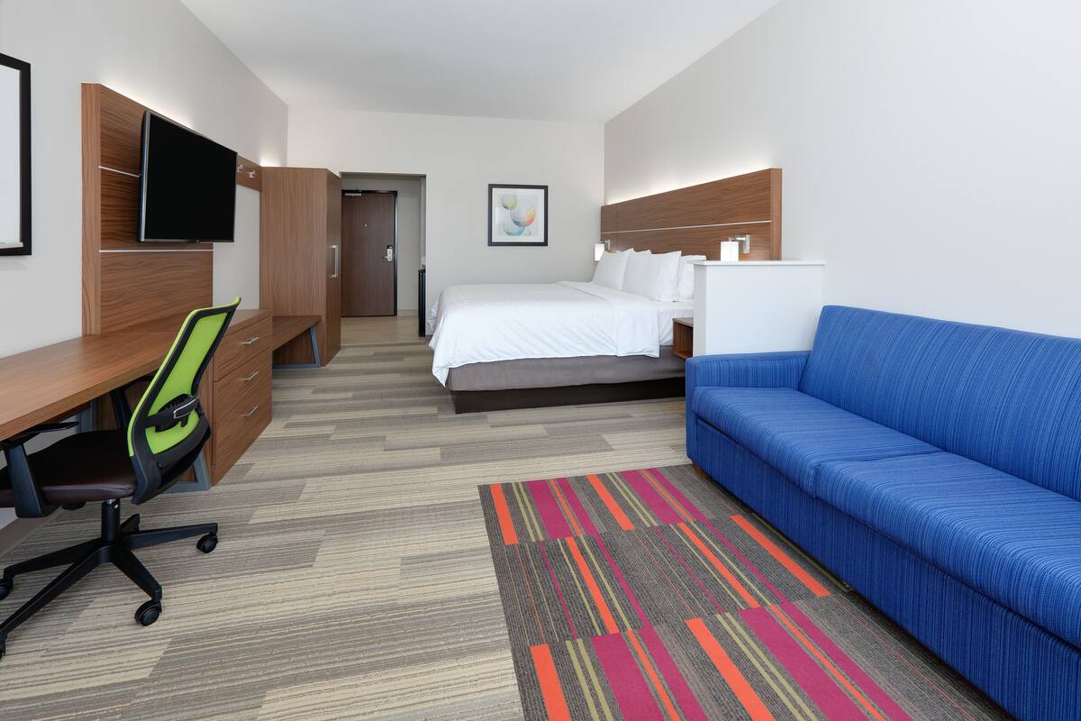 A hotel room with a bed, a blue sofa, a desk with a chair, a TV mounted on a wooden cabinet, and a colorful rug.