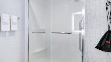 Une salle de bain moderne avec une douche à parois de verre, un porte-serviettes avec deux serviettes blanches et un sac noir suspendu à un crochet.