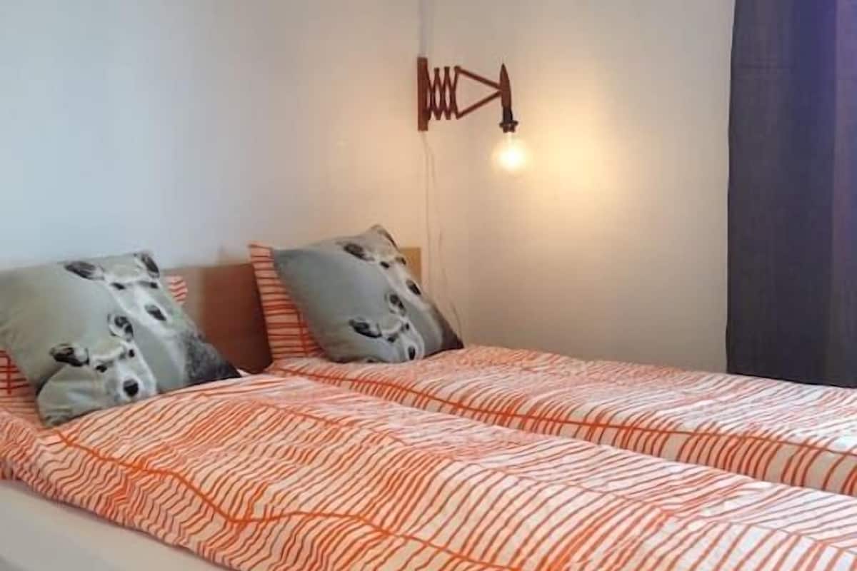 A double bed with an orange and white striped bedspread, two gray pillows with animal prints, and a wall-mounted light fixture.