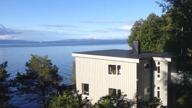 En moderne bygning med to etager, fladt tag, store vinduer og udsigt til en rolig sø og fjerne bjerge.