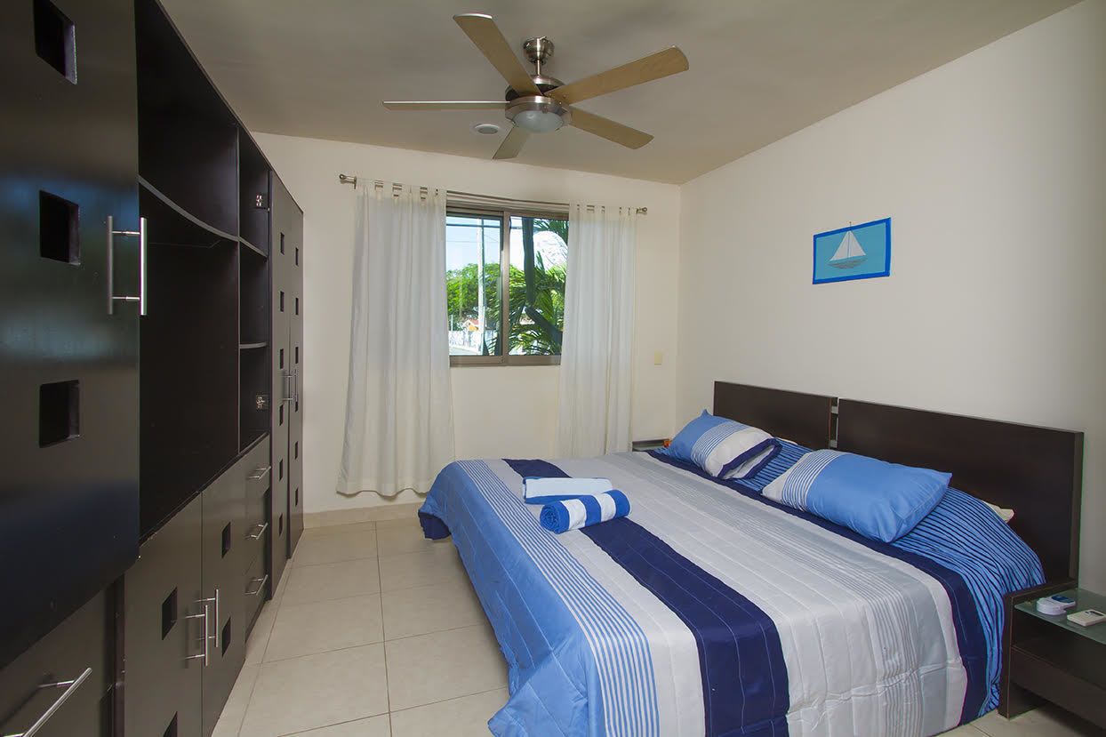 Quarto de hotel com cama, ventoinha no teto, janela com cortinas, mesa de cabeceira e armário.