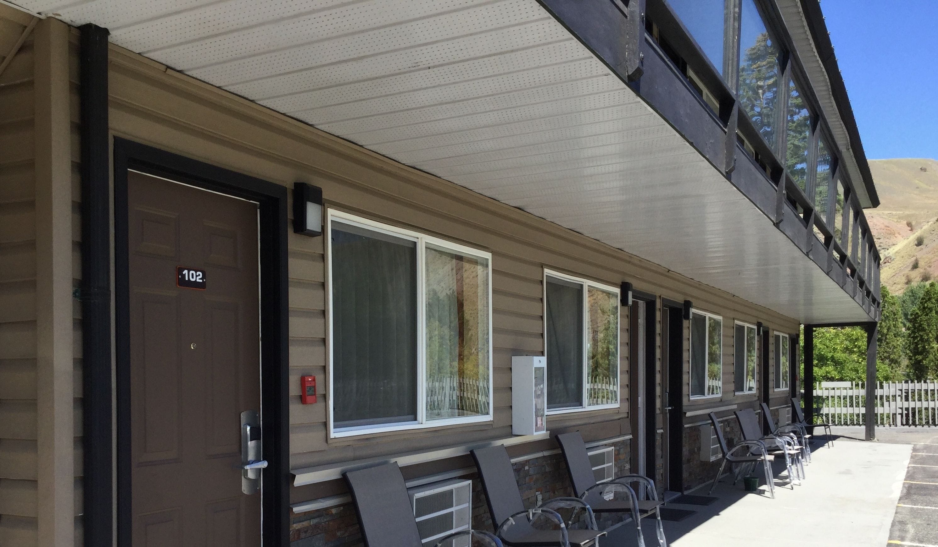 A motel with a row of rooms, each with a door marked with a number. There are outdoor chairs and tables on the porch, and a mountain is visible in the background.