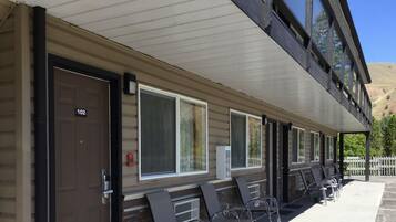 A motel with a row of rooms, each with a door marked with a number. There are outdoor chairs and tables on the porch, and a mountain is visible in the background.