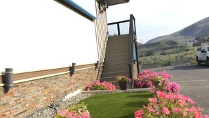 A rooftop garden with a brick wall, potted pink flowers, and a view of a road and hills. - Riverside Motel (Cache Creek)