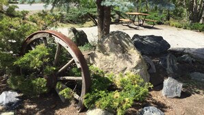 A rustic outdoor setting with a large metal wheel, rocks, and a picnic table in a natural landscape. - Riverside Motel (Cache Creek)