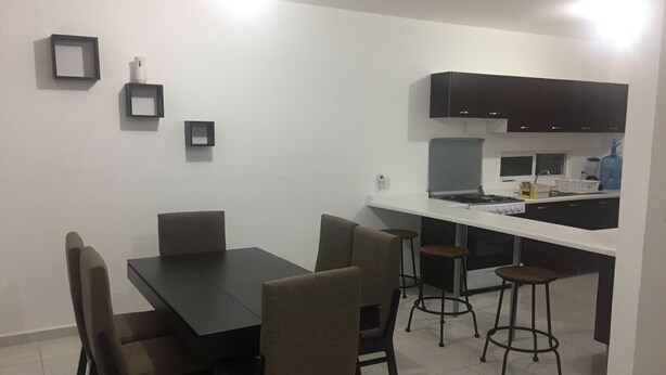 A compact kitchen with a white countertop, black cabinets, and a gas stove. There are four stools and a dining table with chairs.