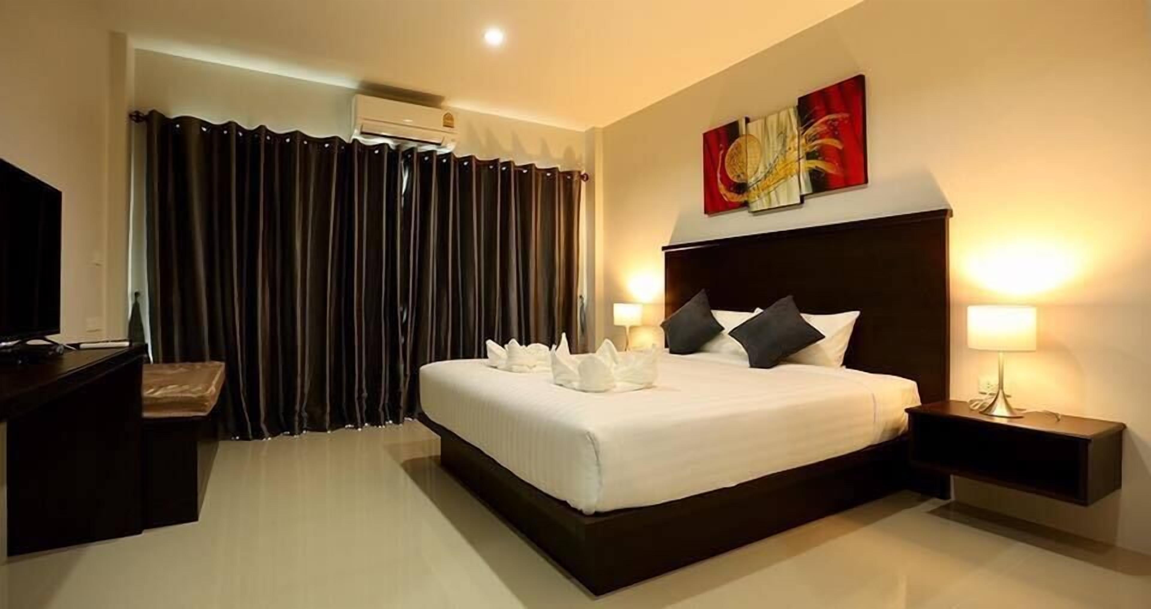 Quarto de hotel com uma cama grande, cortinas escuras, ar condicionado fixado na parede, abajur e uma obra de arte na parede.
