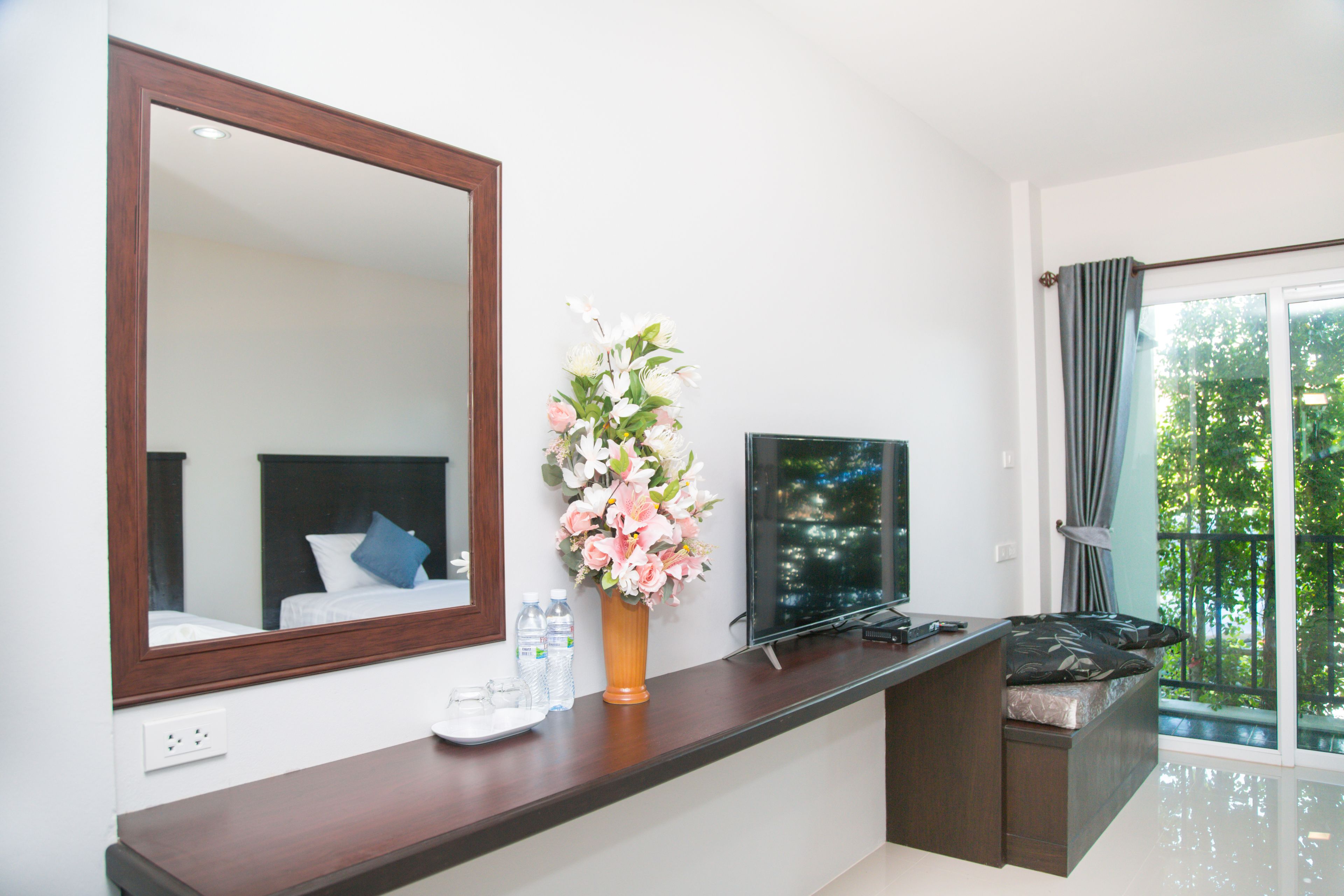 A modern hotel room with a large mirror, a flat-screen TV, a wooden console table, a vase of flowers, and a balcony with a view of greenery.
