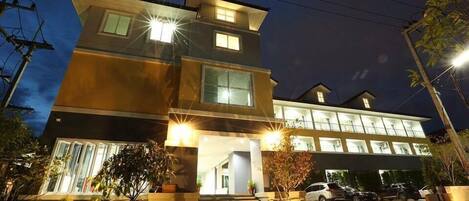 A multi-story hotel with a modern design, featuring a prominent entrance with steps, a covered driveway, and a parking area with several cars.