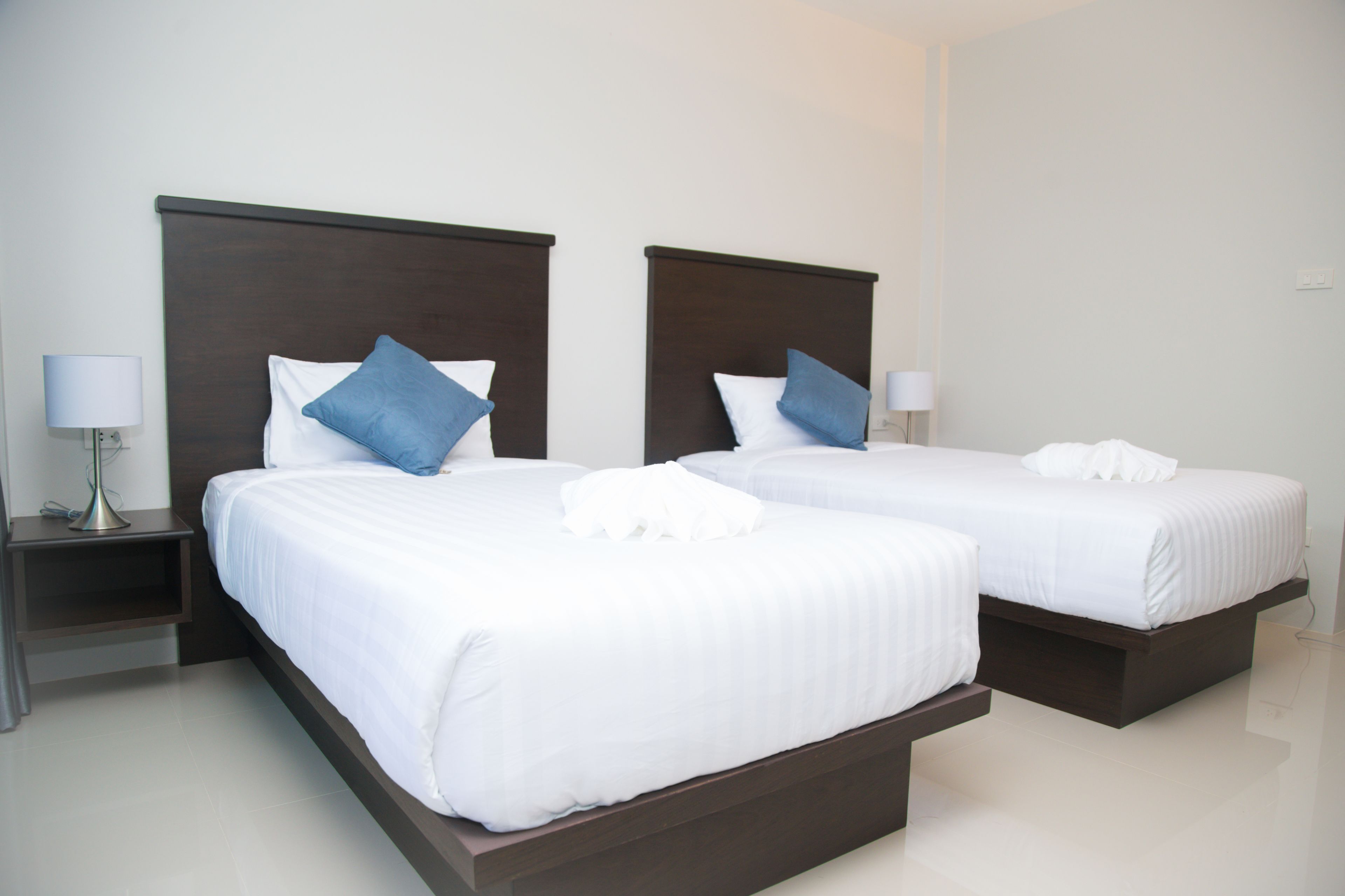 Dos camas individuales con cabeceros oscuros, ropa de cama blanca y almohadas azules. Cada cama tiene una mesita de noche con una lámpara.