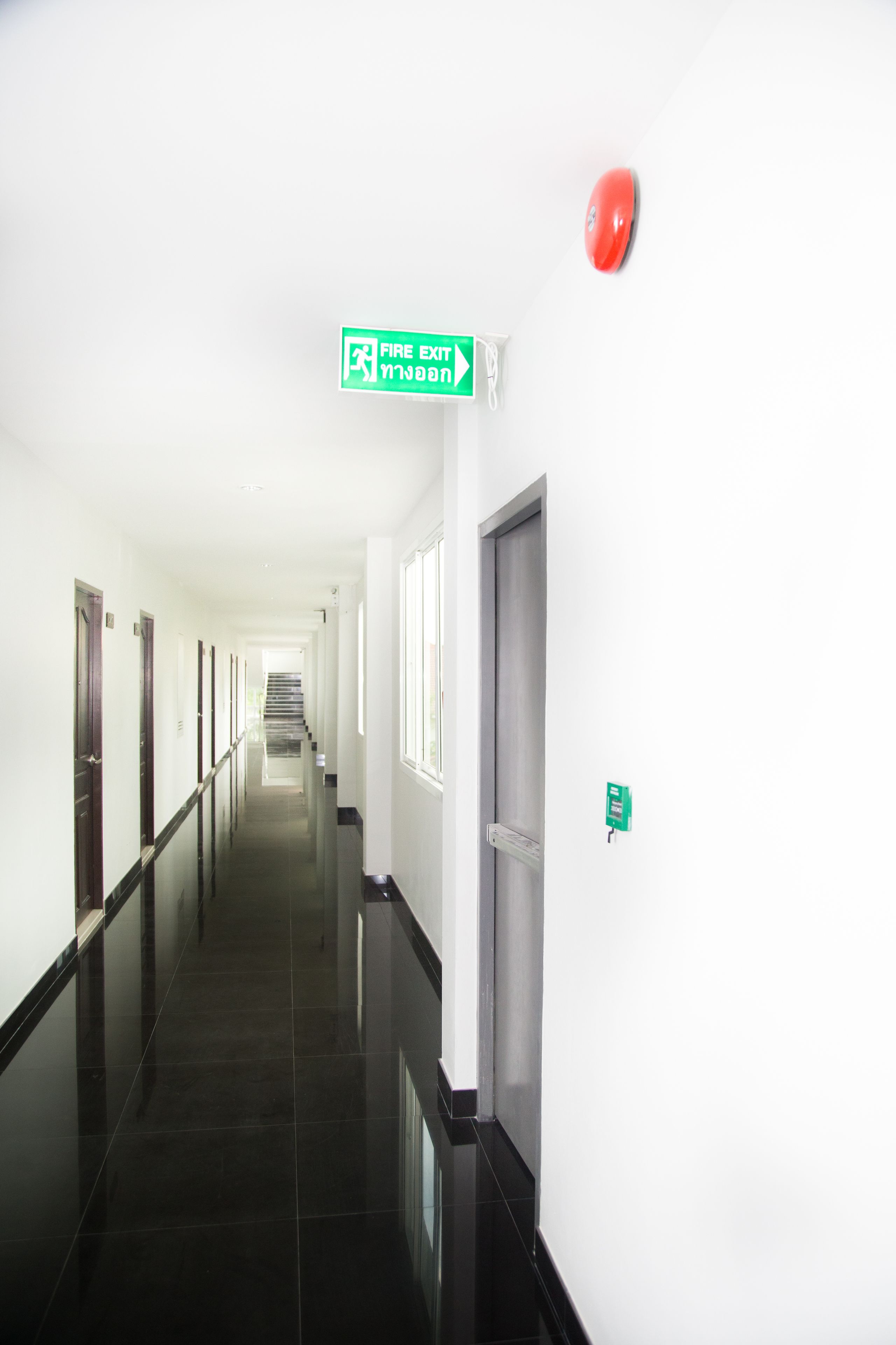 A long, narrow hallway with a fire exit sign, a red emergency alarm, and a door at the end.