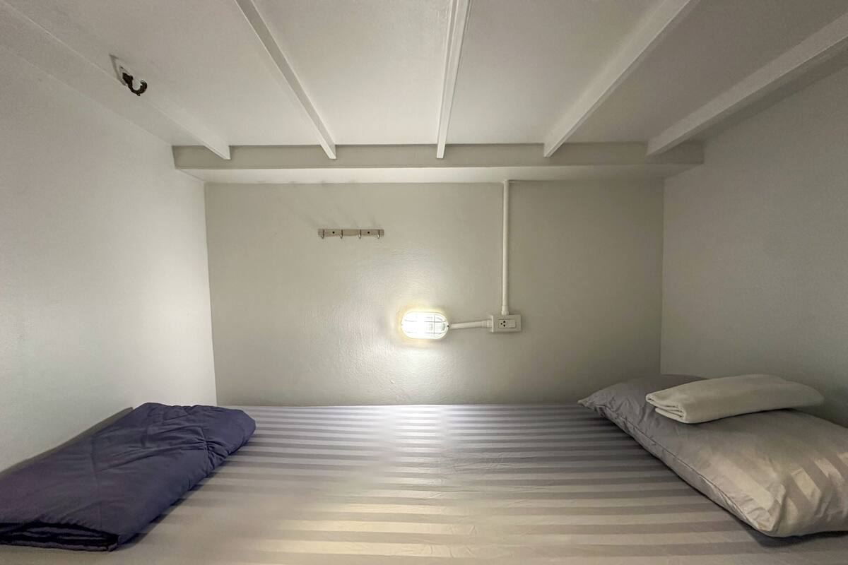 A small, single-bed room with a plain white ceiling, a wall-mounted light, and a power outlet.