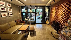 A cozy living area with a leather sofa, a small round table, and a bookshelf. There is a guitar leaning against the wall and a fan in the corner. - 84 Gallery - Hostel (Chiang Mai)