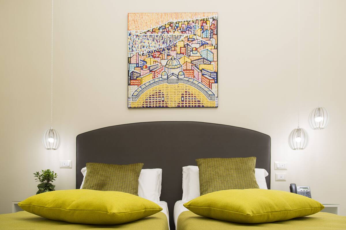 Ett hotellrum med en säng som har två gröna kuddar, en vit kudde och en sänggavel. Ovanför sängen hänger ett färgstarkt konstverk som föreställer en stadsbild.