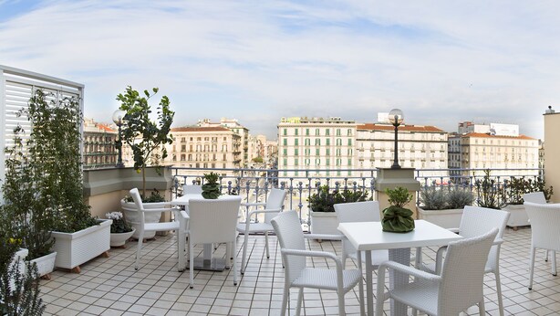 A rooftop terrace with white wicker furniture, potted plants, and a view of city buildings. - The Five Rooms Napoli (Naples)