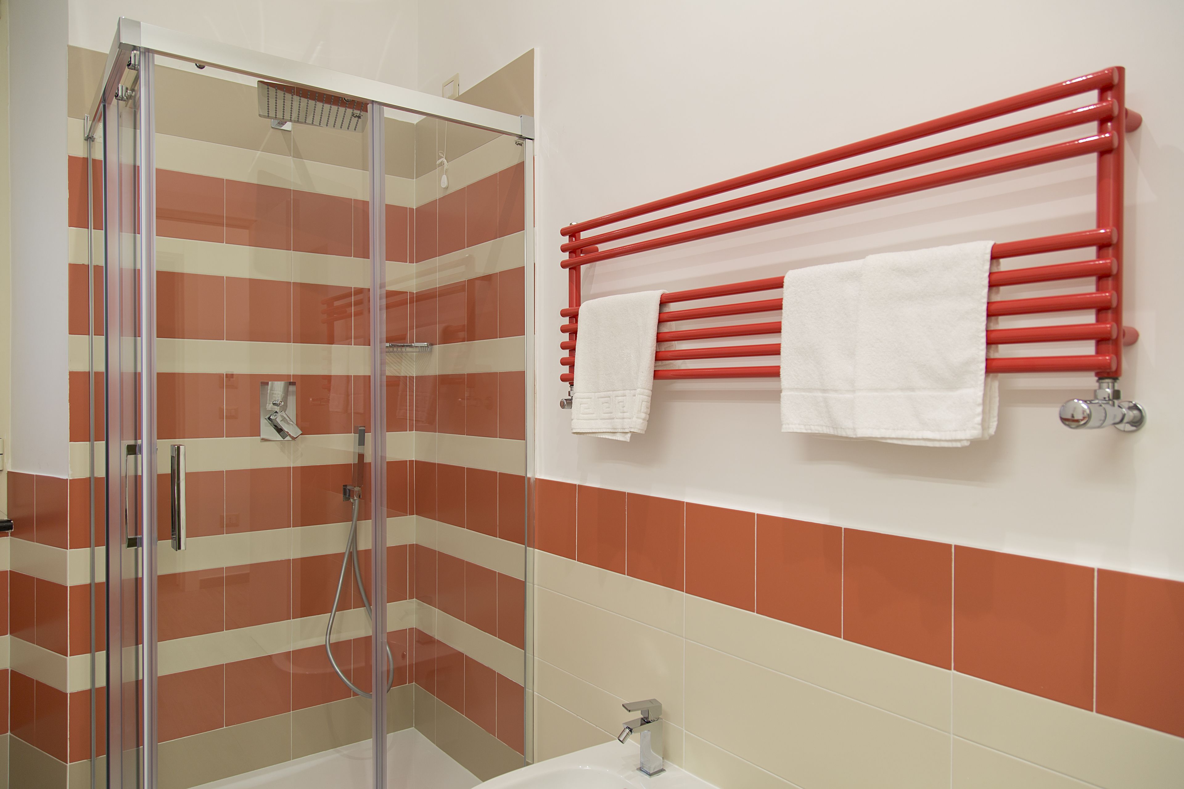 A bathroom with a glass shower enclosure, a red towel rack with two towels, and a white sink.