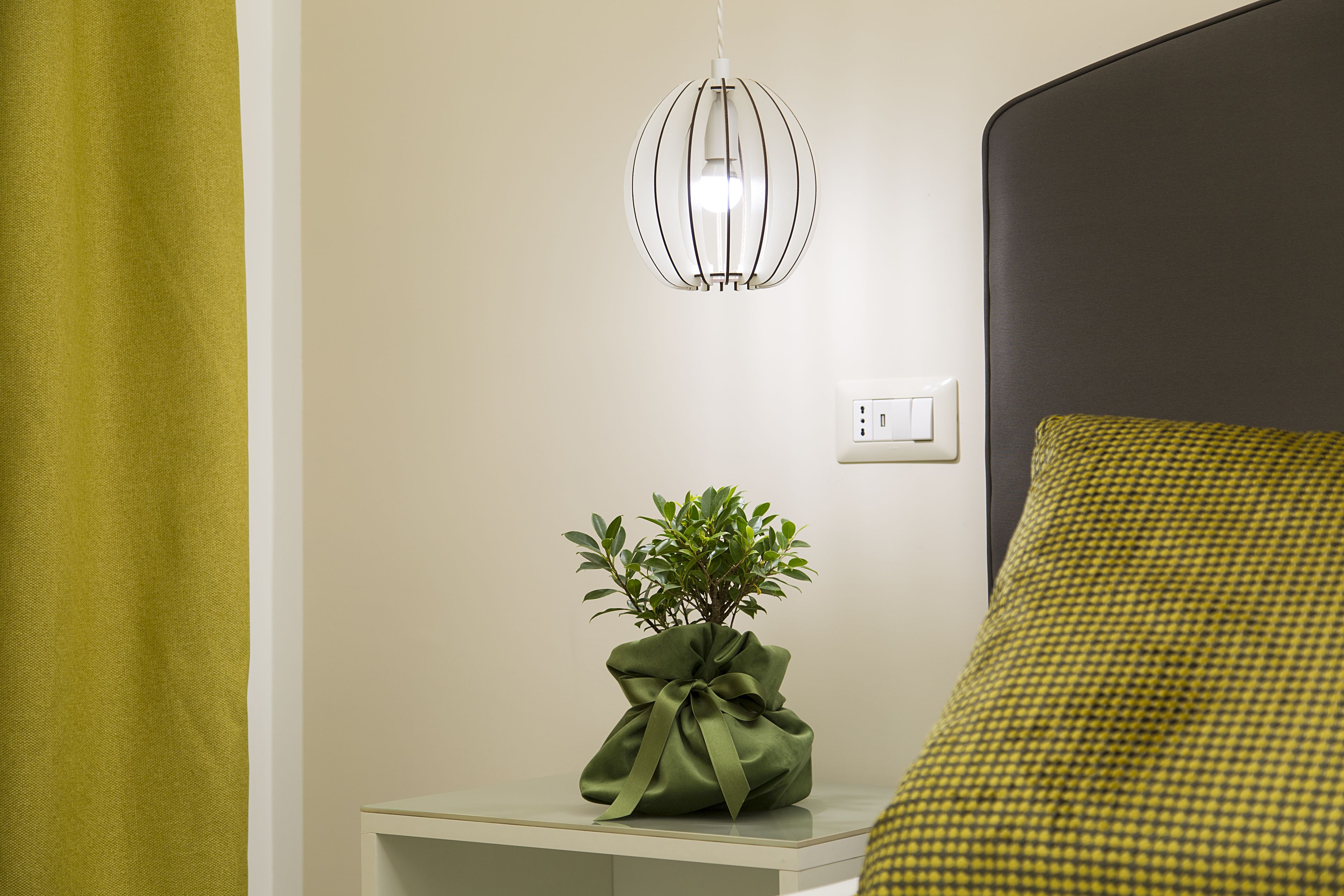 A neatly arranged bedroom with a bed, a bedside table with a green plant, a hanging light fixture, and a power outlet.