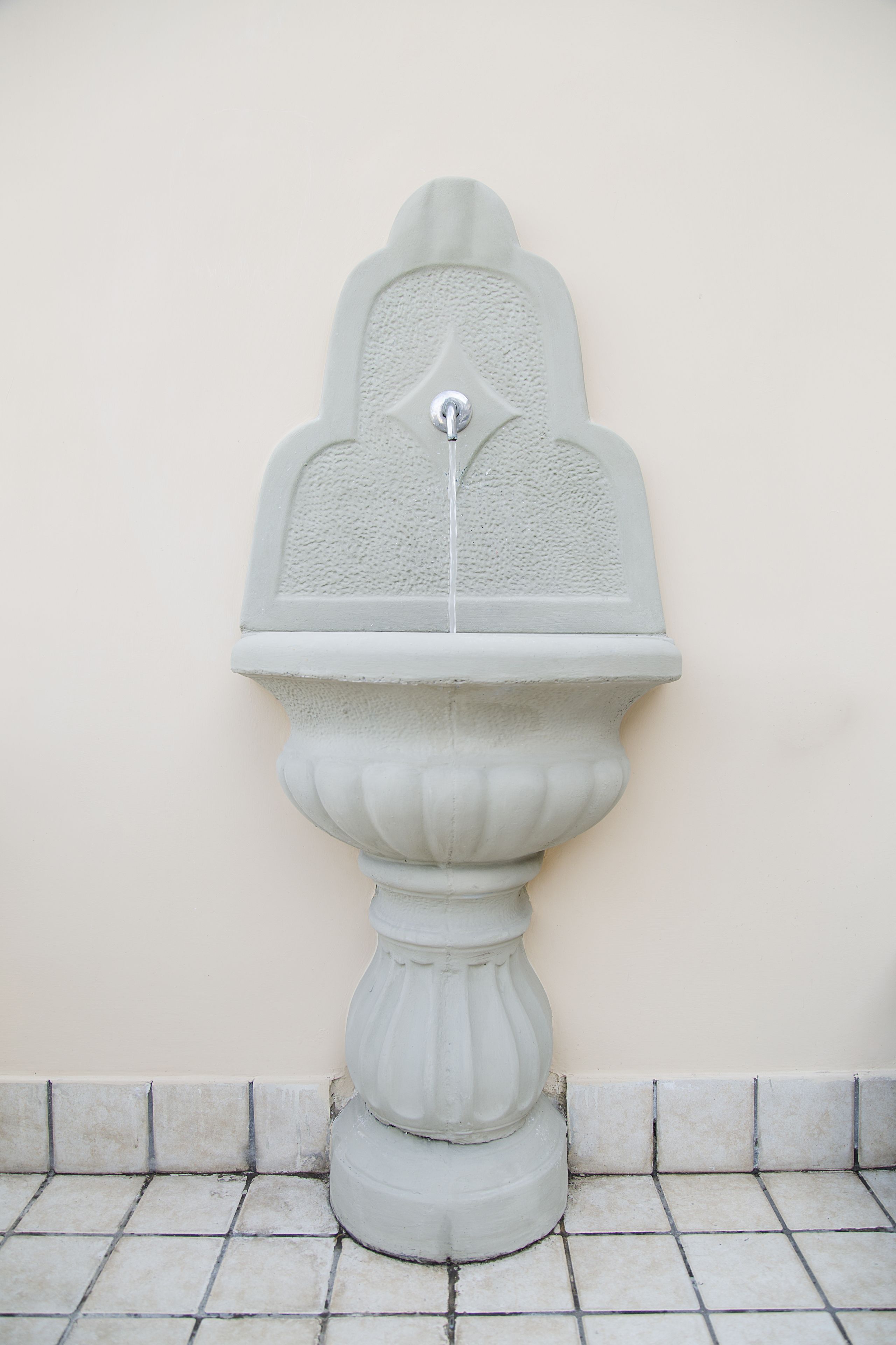 A white, freestanding fountain with a decorative top and a pedestal base, set against a plain wall with a tiled floor.