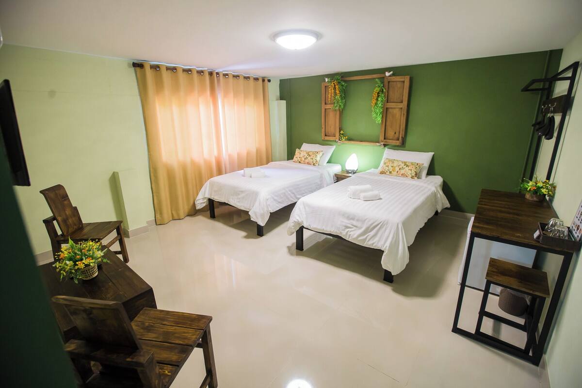 A hotel room with two beds, a wooden desk, a chair, a table with a vase of flowers, and a green wall with a wooden cabinet above the beds.