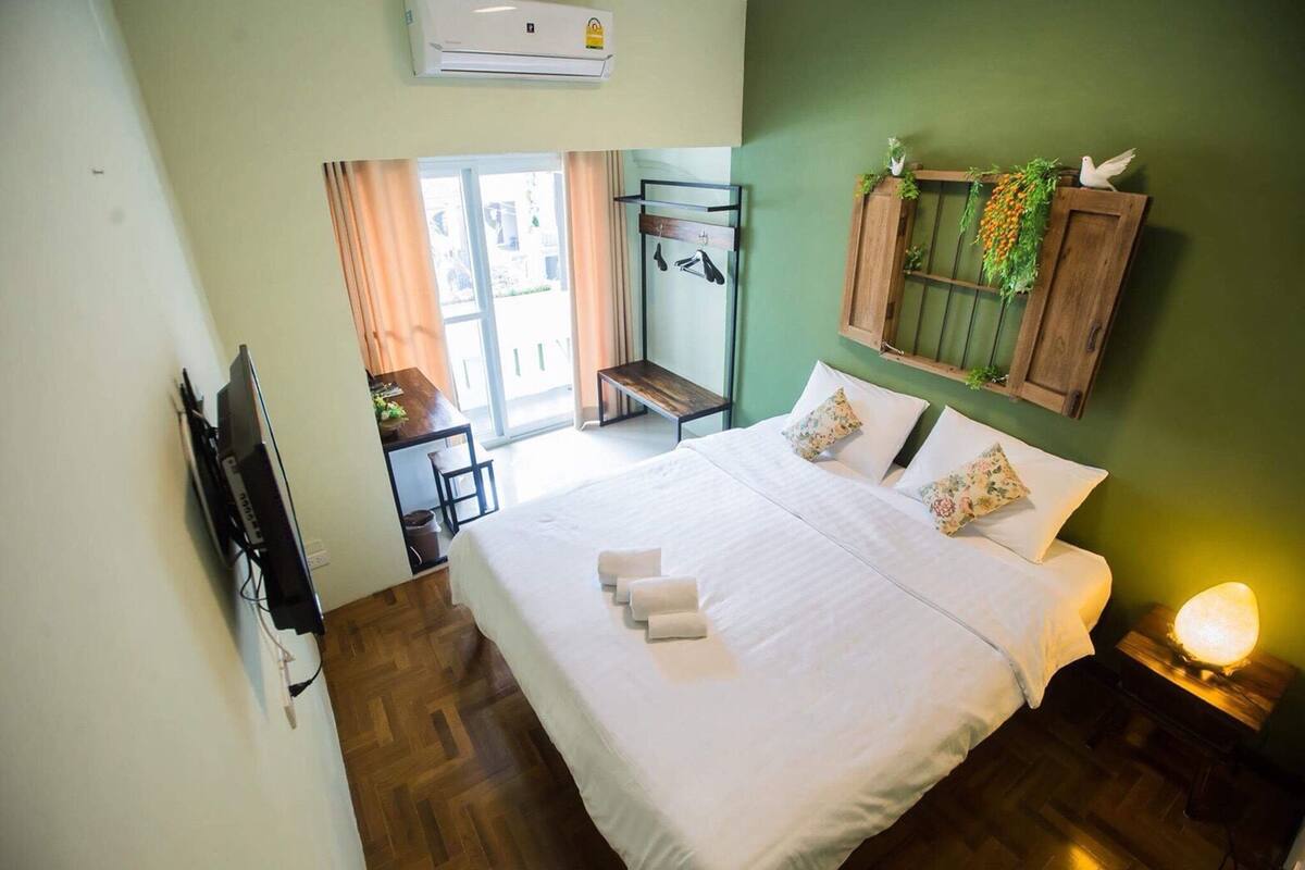 A hotel room with a bed, a wooden bench, a wall-mounted rack, and a nightstand with a lamp.