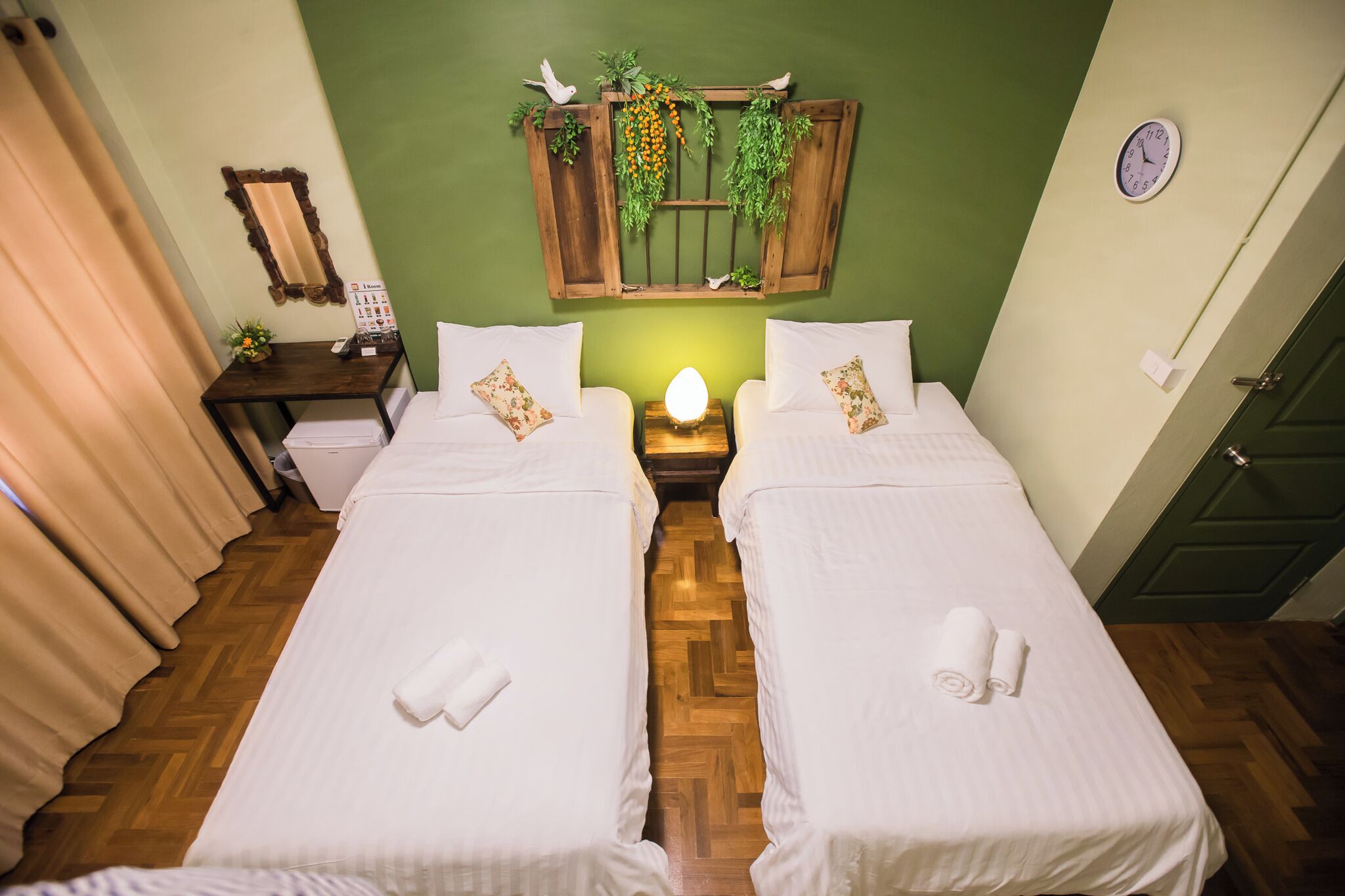 Une chambre d’hôtel avec deux lits, une tête de lit en bois agrémentée de plantes, une horloge et un mur vert.