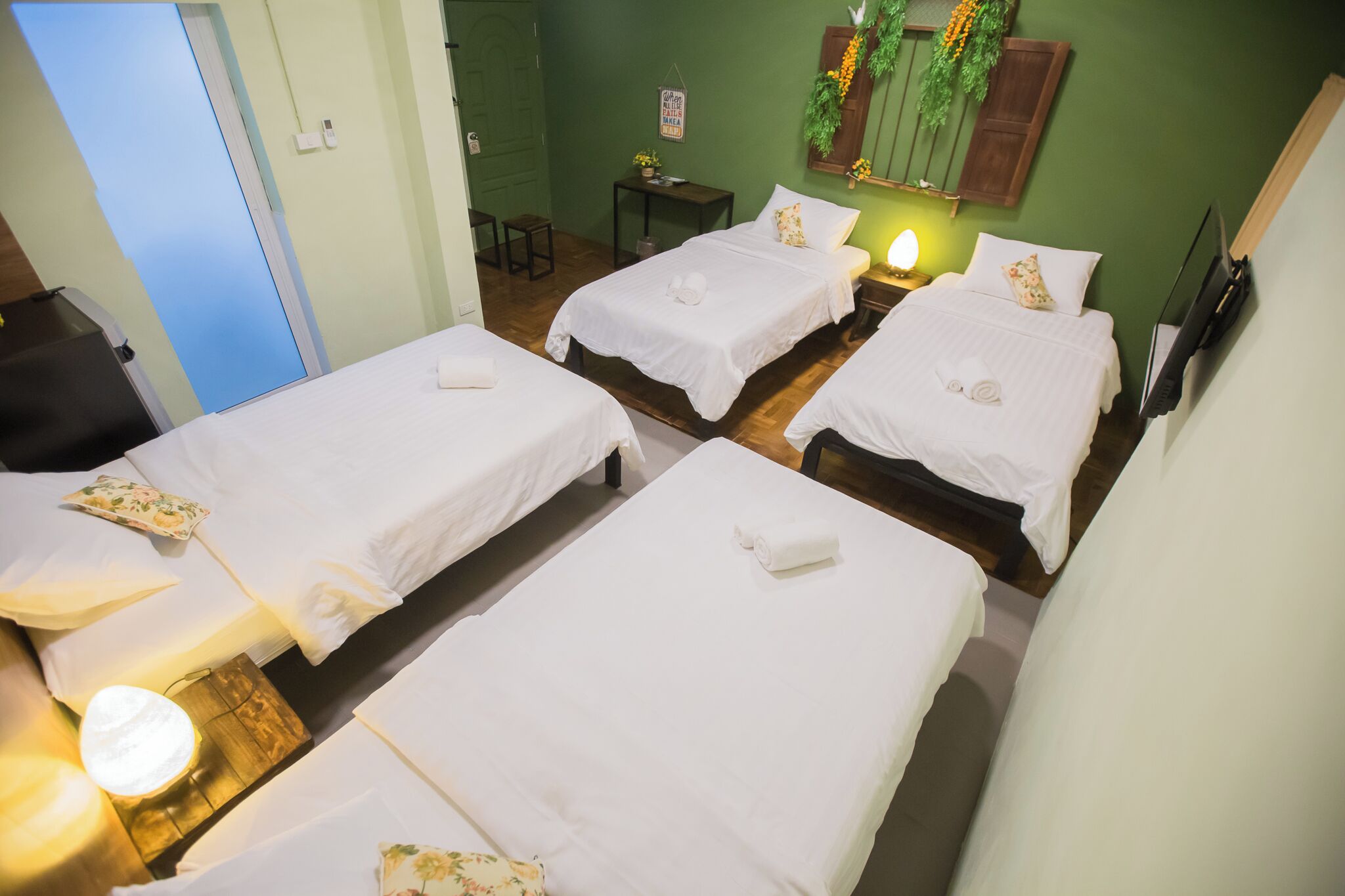 A hotel room with four neatly made beds, a small table with a lamp, a TV mounted on the wall, and a door with a light switch.
