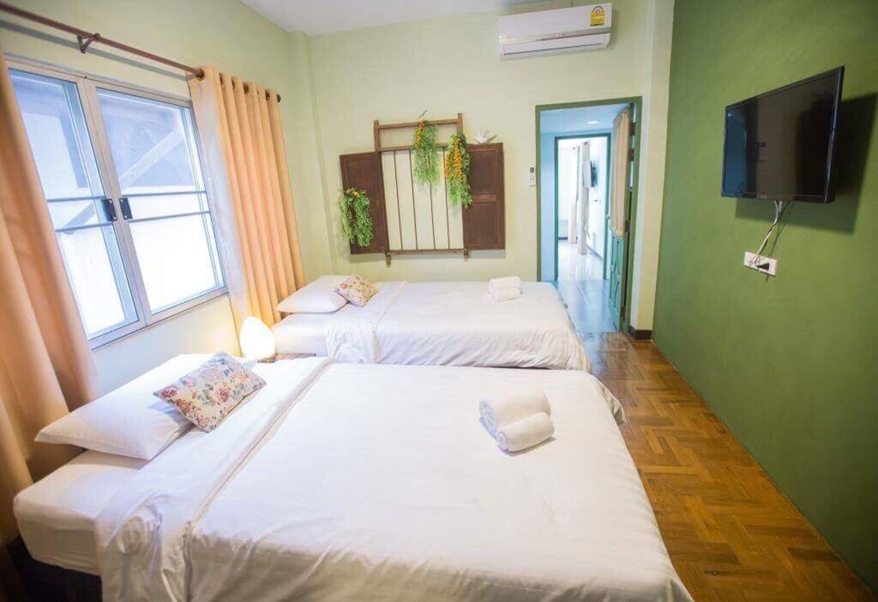 A hotel room with two beds, a window with curtains, a wall-mounted TV, and a door leading to another room.