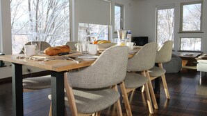 A dining area with a wooden table, grey upholstered chairs, and a large window with a view of snow-covered trees. - Forest Hideaway (Niseko)