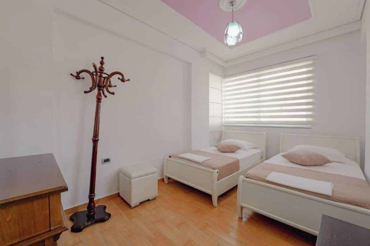 A bedroom with two single beds, a wooden coat stand, a small table, and a window with blinds.